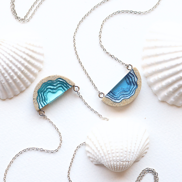 Two dainty beach sand and blue resin necklaces with grey background
